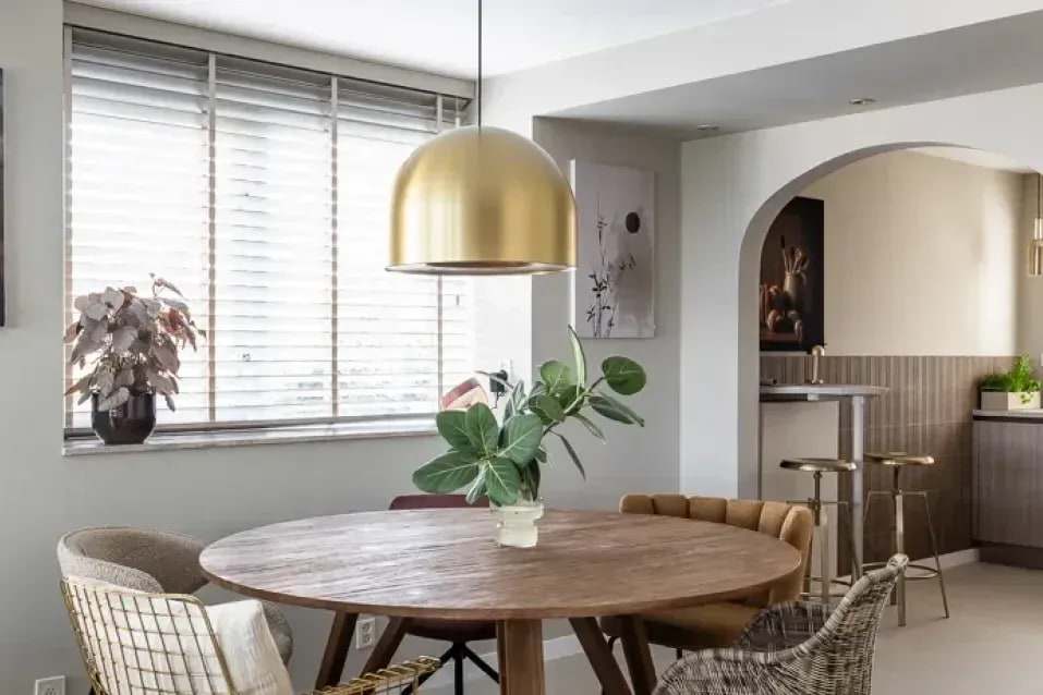 Eethoek met houten jaloezieën voor de ramen, een ronde houten tafel en een gouden hanglamp