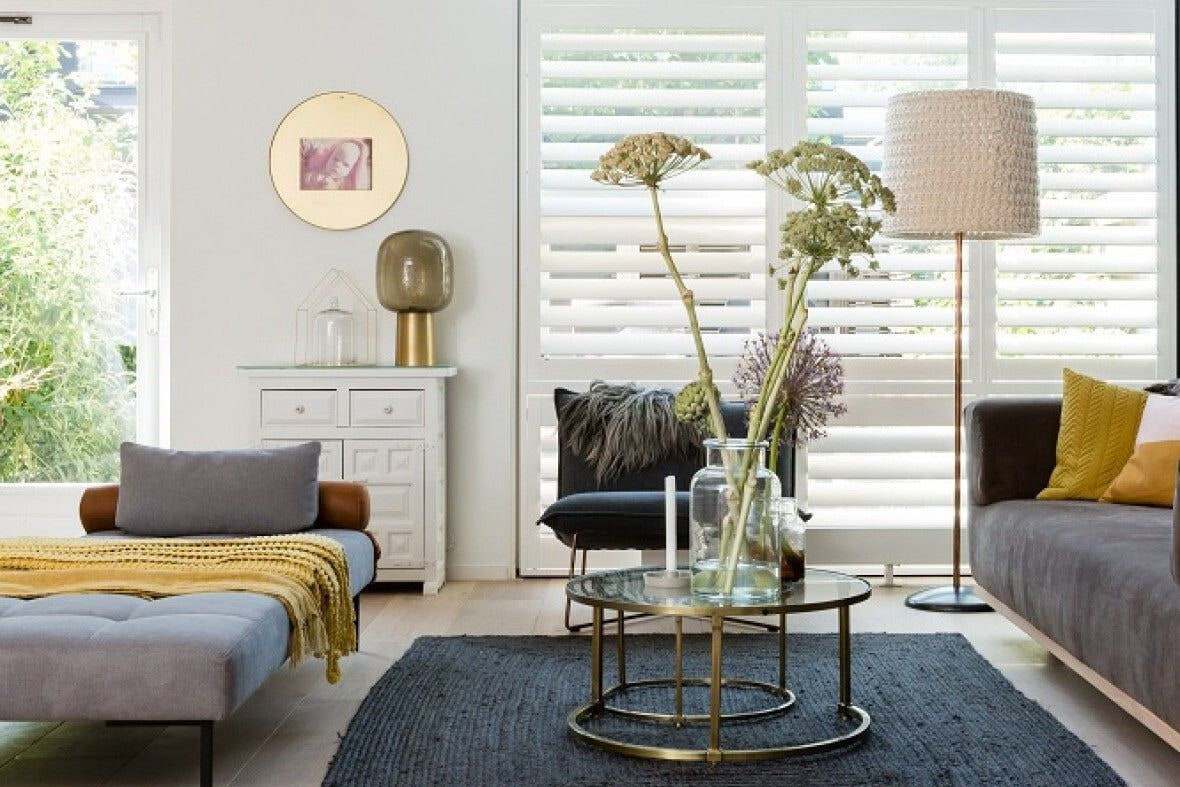 Witte shutters voor een hoog raam in een lichte woonkamer, met een glazen salontafel en een staande lamp